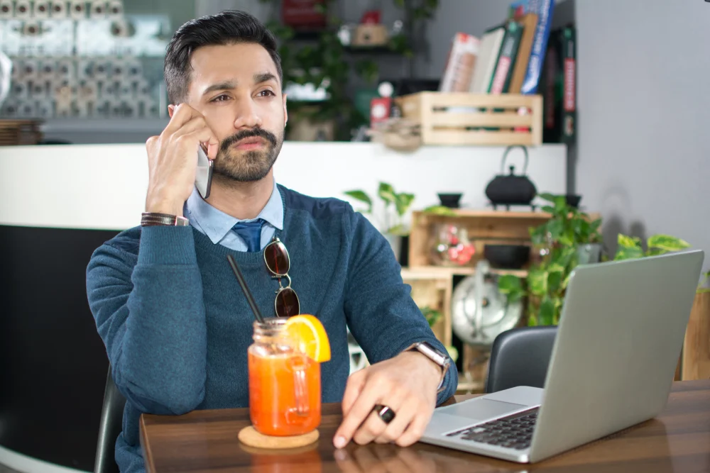 thoughtful indian man listening to cell phone and working on laptop